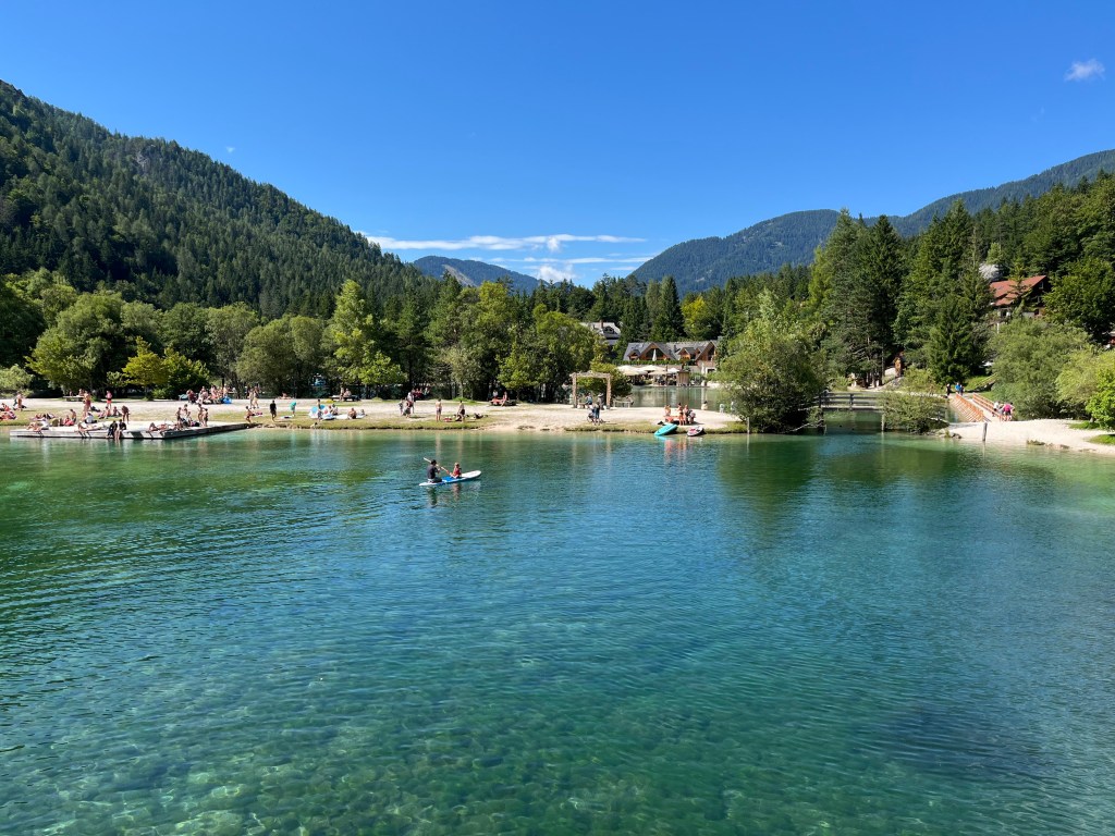 Vista de un lago transparente rodeado de montañas y árboles, con personas tomando el sol y disfrutando de actividades acuáticas.