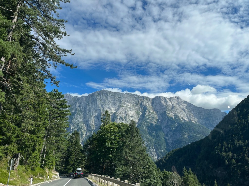 Carro circulando por una carretera rodeada de árboles, con montañas majestuosas y nubes en el cielo.