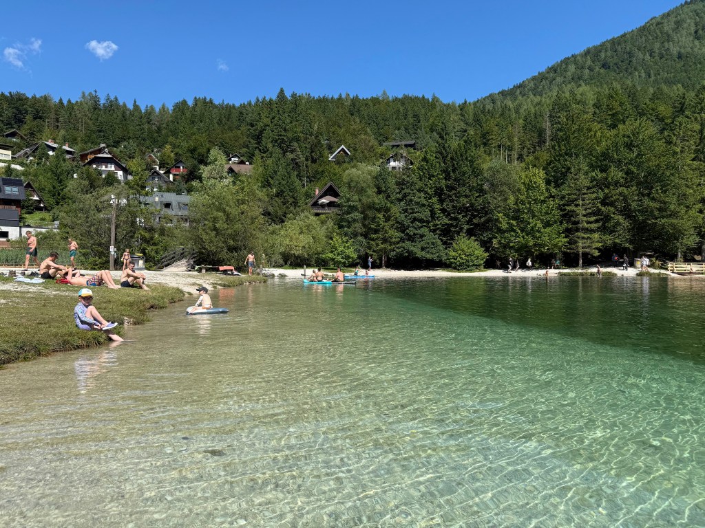 Un lago claro y tranquilo rodeado de vegetación, con personas disfrutando del sol y la natación en una playa de arena.
