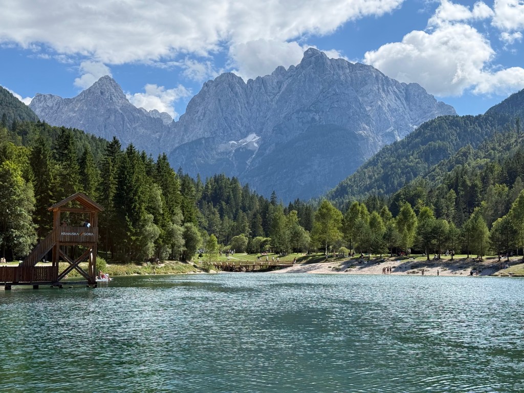 Lago rodeado de montañas con vegetación abundante y un cielo azul con nubes. Se observa una estructura de madera y personas disfrutando de la naturaleza.